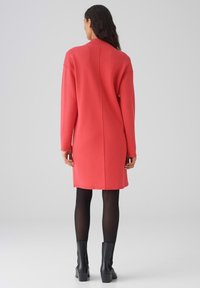 Abito corallo oversize con collo alto, maniche lunghe e taglio dritto. Realizzato in materiale morbido, presenta una cucitura centrale sul retro.