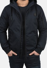 Veste zippée bleu marine avec capuche, dotée de poches latérales, d'une poche sur la manche gauche et d'un logo circulaire sur la poitrine.