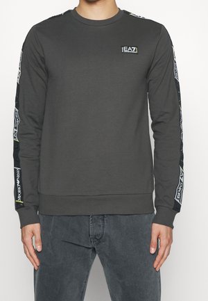 Mężczyzna noszący ciemnoszarą bluzę z długim rękawem z logo EA7 Emporio Armani na piersi i markowymi paskami na rękawach.