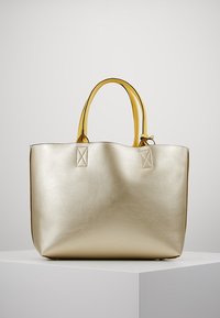 Grand sac cabas métallique doré avec deux poignées jaunes, exposé sur un piédestal blanc sur fond uni.
