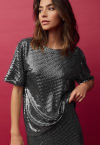 Top nero con paillettes, a maniche corte e taglio largo, caratterizzato da una texture lucida. Lo sfondo è di un colore rosa uniforme.