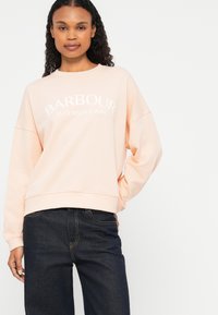 Heledat roosa spordipusa, millel on valge tekst "BARBOUR INTERNATIONAL", mugav lõige, soonikkoes mansetid, koos tumedate teksadega.