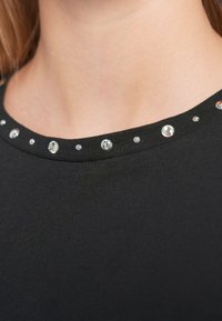 Μαύρο μπλουζάκι με στρογγυλή λαιμόκοψη διακοσμημένη με κυκλικές, λαμπερές λεπτομέρειες γύρω από την άκρη, κατασκευασμένο από μαλακό, ελαστικό υλικό.
