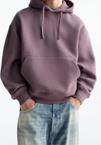 Felpa a pullover oversize in viola tenue con tasca frontale, polsini a coste e cappuccio con cordino, abbinata a jeans scoloriti azzurri chiari.