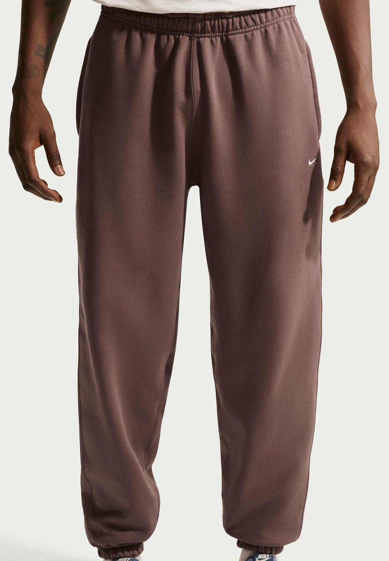 Persoon draagt losse bruine Nike-joggingbroek met elastische boorden en tailleband, handen langs het lichaam, wit shirt gedeeltelijk zichtbaar bij de taille.