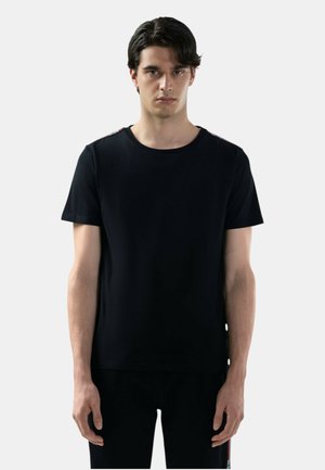 Giovane uomo con capelli scuri che indossa una T-shirt nera a maniche corte semplice e pantaloni neri abbinati, in piedi davanti a uno sfondo grigio chiaro.