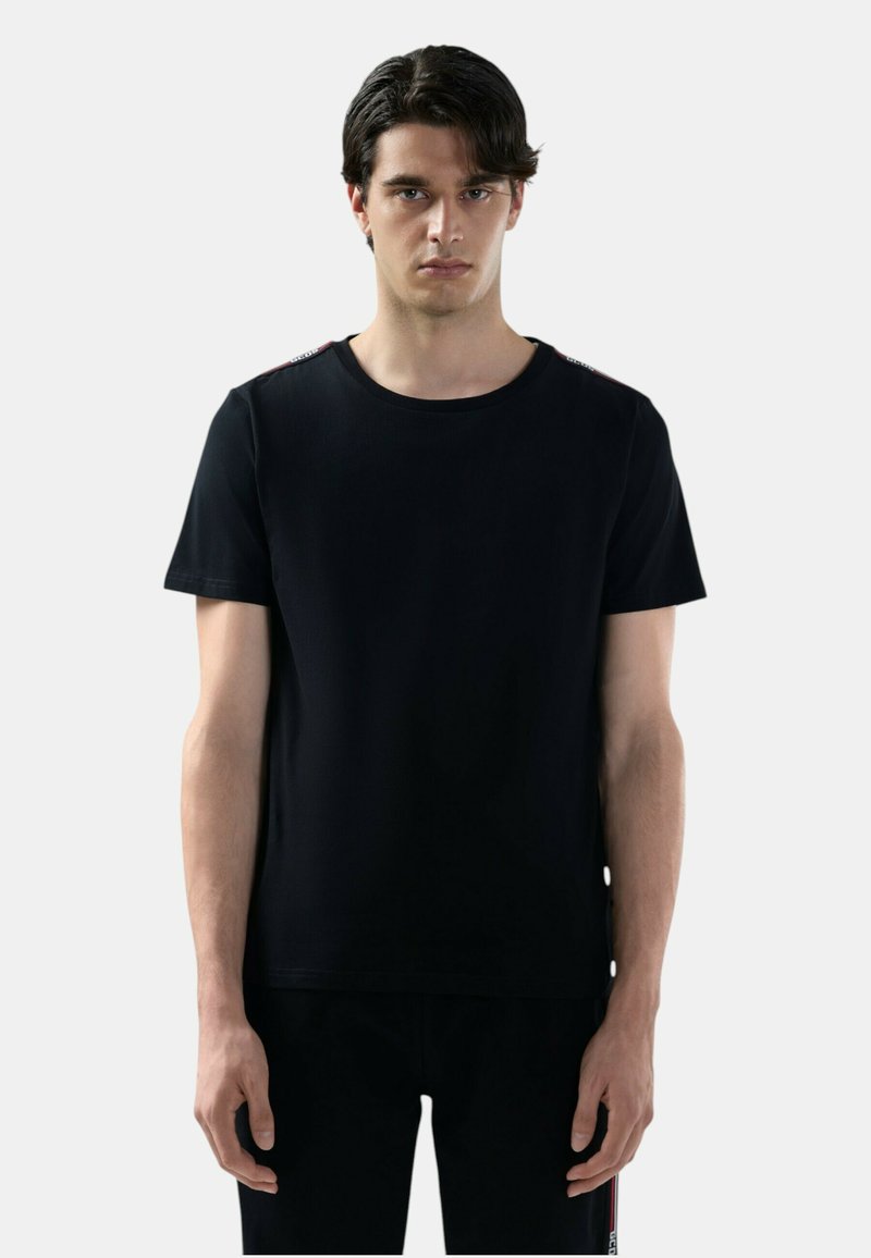 Giovane uomo con capelli scuri che indossa una T-shirt nera a maniche corte semplice e pantaloni neri abbinati, in piedi davanti a uno sfondo grigio chiaro.