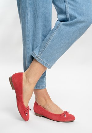 Feet wearing red suede ballet flats with bows and pearls, paired with cropped light blue jeans against a plain background.
