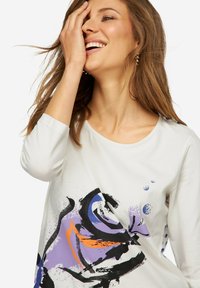 Weißes langärmliges Oberteil mit rundem Ausschnitt, das abstrakte Muster in Lila, Schwarz und Orange aufweist. Glatte Stoffoberfläche, lässiges Design.
