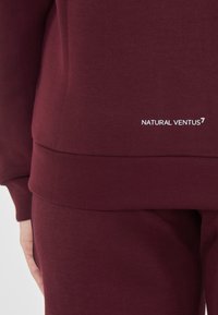 Burgundowa bluza z gładką fakturą, elastycznym pasem i białym napisem "NATURAL VENTUS" w pobliżu brzegu.