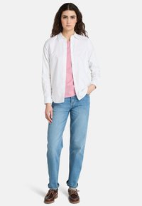 Camisa blanca de botones sobre una camiseta rosa, combinada con jeans azul claro y zapatos marrones, con un ajuste relajado y diseño clásico.