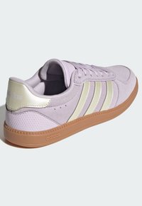 Fioletowy zamszowy sneaker z beżowymi syntetycznymi paskami, okrągłym noskiem, płaską gumową podeszwą oraz teksturowaną tylną częścią. Posiada oznaczenie „adidas”.