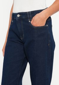 Jeans din denim albastru închis cu o textură netedă, buzunare frontale și un sistem de închidere cu un buton argintiu. Cusăturile vizibile accentuează cusăturile.