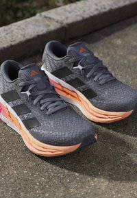 Paire de chaussures de course grises et noires avec des semelles dégradées orange reposant sur le trottoir près d'un bord en pierre.