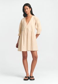 Kurzes, cremefarbenes Kleid aus leichtem Stoff. Verfügt über puffärmel, einen tiefen V-Ausschnitt und Knöpfe an der Vorderseite.