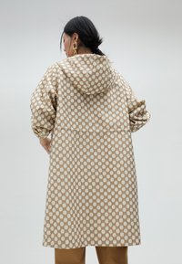 Długa beżowa kurtka z geometrycznym wzorem w kwadraty i diamenty. Posiada kaptur, mankiety oraz luźny krój.