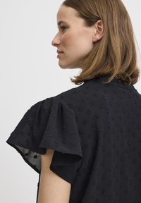 Blusa nera di tessuto strutturato con maniche a sbuffo, motivo a pois e scollatura rotonda, che mette in risalto una finitura di tessuto liscia e una silhouette rilassata.