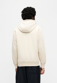 Μπεζ φούτερ με κουκούλα και άνετη εφαρμογή, με ριμπ μανσέτες και τελείωμα, με ομαλή υφή. Όψη από πίσω, φορεμένο με φαρδιά μαύρα παντελόνια.