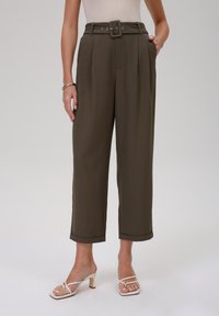 Pantalon taille haute vert olive à jambes larges, avec plis devant, ceinture en tissu et revers roulés. Associé à des sandales blanches à brides.