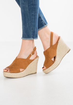Pieds en jean bleu à ourlet effiloché portant des sandales compensées en daim beige avec des lanières croisées et des talons espadrilles tressés sur un sol blanc.