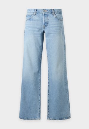 Femme aux longs cheveux foncés portant un haut gris sans manches, un jean bleu clair à jambes larges et des talons ouverts noirs, debout avec une main dans la poche.