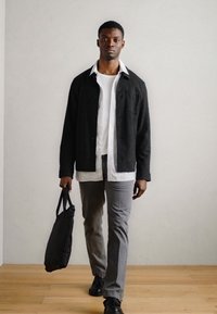 Veste noire texturée sur une chemise blanche, accompagnée de pantalons gris et de chaussures noires. La personne porte un sac fourre-tout noir. Fond en bois.