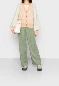 Dzianinowy kardigan w kolorze beżowym i miętowym z dekoltem w serek i guzikami, zestawiony z szerokimi zielonymi spodniami i jasnobeżową torebką. Masywne buty.
