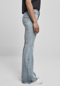 Ljust blå utsvängda jeans med en slät textur, fem fickor och medelhög midja. Kombinerade med vita sneakers, sett från sidan.
