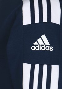 Tessuto blu navy con il logo Adidas bianco. Strisce bianche che corrono verticalmente sui lati, aggiungendo contrasto al materiale scuro.