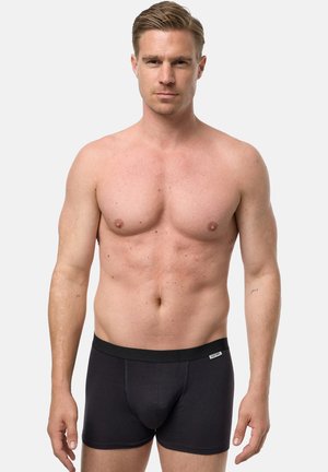 Gespierde, shirtloze man met een lichte huid en kort blond haar, gekleed in zwarte boxershorts, staand tegen een effen lichte achtergrond.