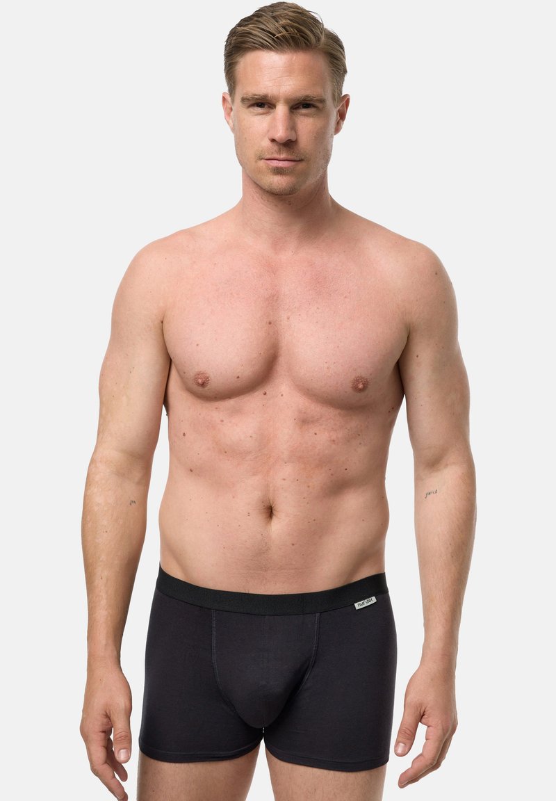 Umięśniony, nagi mężczyzna o jasnej karnacji i krótkich blond włosach, ubrany w czarne bokserki, stoi na tle jasnego, jednolitego tła.