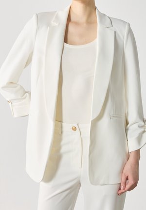 Kobieta ubrana w biały żakiet z podwiniętymi mankietami, dopasowane białe spodnie i biały top, stojąca na jednolitym tle.