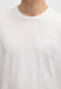 Bílý bavlněný tričko s kulatým výstřihem, se malým, texturovaným bílým čtvercovým logem se nápisem "BOSS". Měkká textura a lehký design.