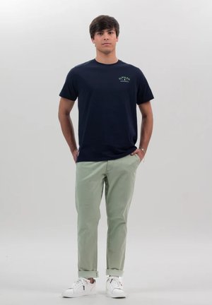 Joven con las manos en los bolsillos, viste una camiseta azul marino, pantalones verde claro remangados en los bajos y zapatillas blancas sobre un fondo liso.