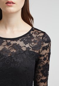 Svart spetsblus med långa ärmar som har blommönster, rund halsringning och ett texturerat nedre panel i ett mjukare material.