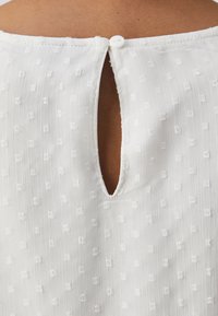 Blusa blanca con un patrón texturizado de pequeños cuadrados, que presenta un diseño de espalda con forma de clave y un cierre con un solo botón blanco.
