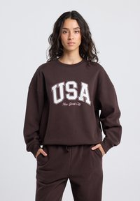 Brown oversized sweatshirt with "USA" and "New York City" printed in white. Features ribbed crew neckline and relaxed fit. Soft fabric texture.