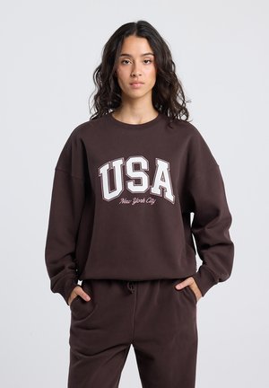 Bruine oversized sweatshirt met "USA" en "New York City" gedrukt in het wit. Heeft een geribbelde ronde hals en een relaxte pasvorm. Zachte stoftextuur.