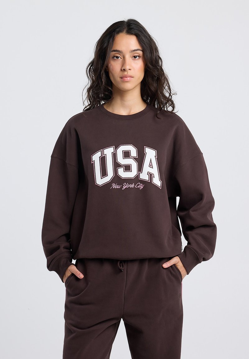 Brown oversized sweatshirt with "USA" and "New York City" printed in white. Features ribbed crew neckline and relaxed fit. Soft fabric texture.