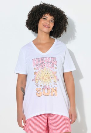 Lachend persoon met krullend haar, draagt een wit V-hals T-shirt met de tekst "Here Comes the Sun" en een zonafbeelding, gecombineerd met rood-wit gestreepte korte broek.
