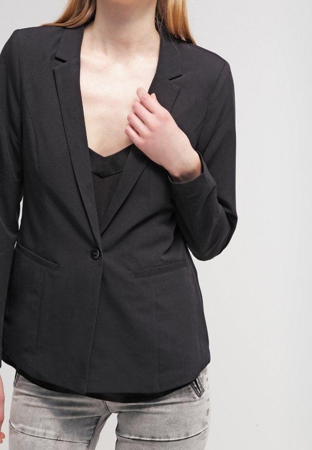 Žena v černém saku přes černý top, která pravou rukou sahá na límec saka, stojící na pozadí v jasné světle.