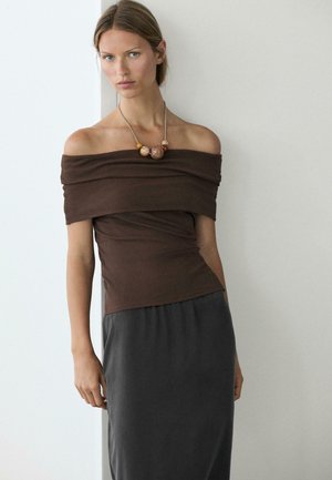 Vrouw draagt een bruin off-shoulder top, een donkere rok en een opvallende kralenketting, staand tegen een effen witte muur.