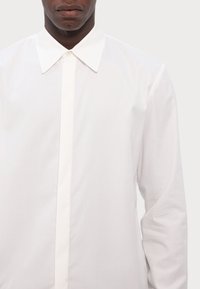 Λευκό πουκάμισο με μακριά μανίκια και μυτερό γιακά, με κλείσιμο μπροστά με κουμπιά και λεία υφή. Απλός σχεδιασμός, χωρίς μοτίβα ή διακοσμητικά στοιχεία.