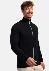 Sudadera negra con cremallera, con textura de espiga, cuello alto, puños acanalados y dobladillo. Presenta una cremallera blanca en contraste.