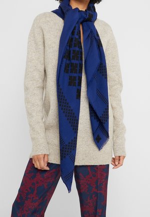 Personne portant un pull en maille beige, une écharpe bleue à motifs nouée autour du cou, et un pantalon aux motifs abstraits rouges et bleu marine, debout devant un fond gris.