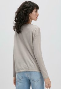 Helles graues Langarmshirt mit lockerem Schnitt, weichem Material, rundem Halsauschnitt und dezenten seitlichen Schlitzen, kombiniert mit hellblauen Jeans.