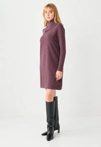 Robe jusqu'aux genoux à manches longues en maille bordeaux foncé avec un col cheminée. Portée avec des bottes noires hautes à bout pointu. Silhouette simple et droite.