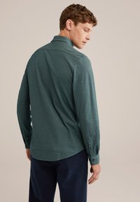 Bărbat întors lateral, îmbrăcat cu o cămașă cu manșete lungi de culoare teal și pantaloni negri închis, pe un fundal simplu bej.