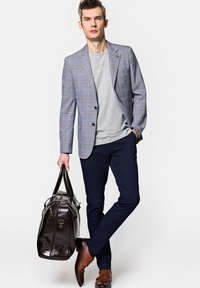 Marynarka w niebieską kratę, szary t-shirt, granatowe spodnie, brązowe skórzane buty i ciemnobrązowa skórzana torba. Gładkie faktury i dopasowany krój.