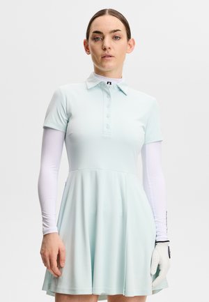 Vrouw met een lichtblauwe jurk met korte mouwen over een witte blouse met lange mouwen, met een golfhandschoen aan haar linkerhand, tegen een effen achtergrond.
